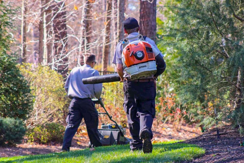 Two mean wearing work uniforms, one with a push lawnmower and the other with a backpack leaf blower working